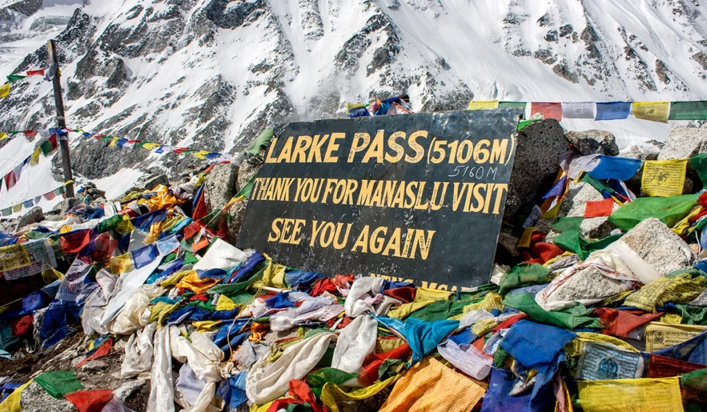 Larkya La Pass welcome board standing at a snowy high-altitude point with prayer flags fluttering, marking the highest point of the Manaslu Circuit Trek.
